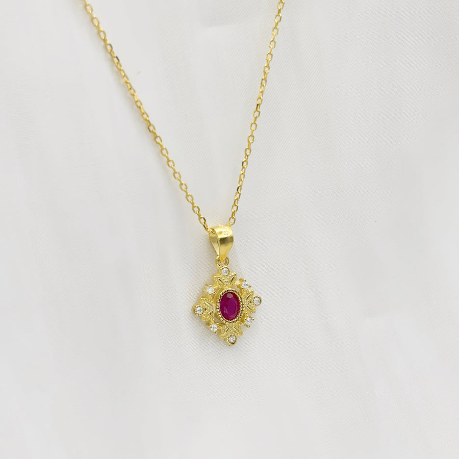 This delicate gold necklace features an intricately designed pendant with a central red cubic zircon, surrounded by a floral pattern and small clear cubic zircon. The elegant design adds a touch of sophistication and charm to any outfit. Perfect for special occasions or everyday wear, this necklace adds a touch of timeless sophistication to any outfit with its brilliant sparkle.