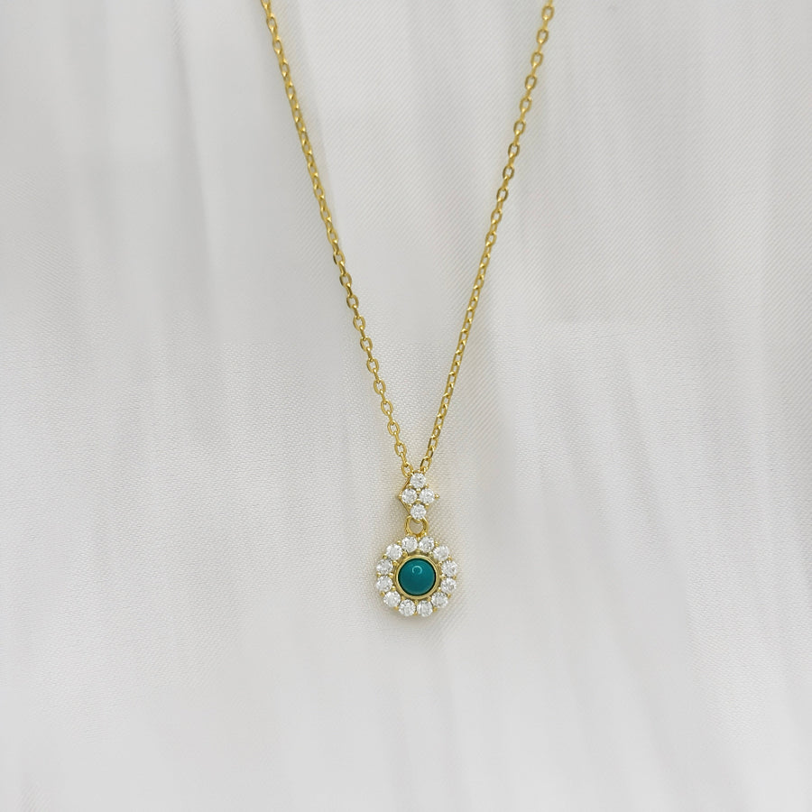 This elegant necklace features a delicate gold chain and a pendant adorned with a central turquoise stone, surrounded by a halo of sparkling cubic zircon. The combination of the vibrant turquoise and the shimmering cubic zircon creates a striking and timeless piece of jewelry. Perfect for any occasion, this necklace adds a touch of sophistication and color to your look, making it a versatile addition to any jewelry collection.