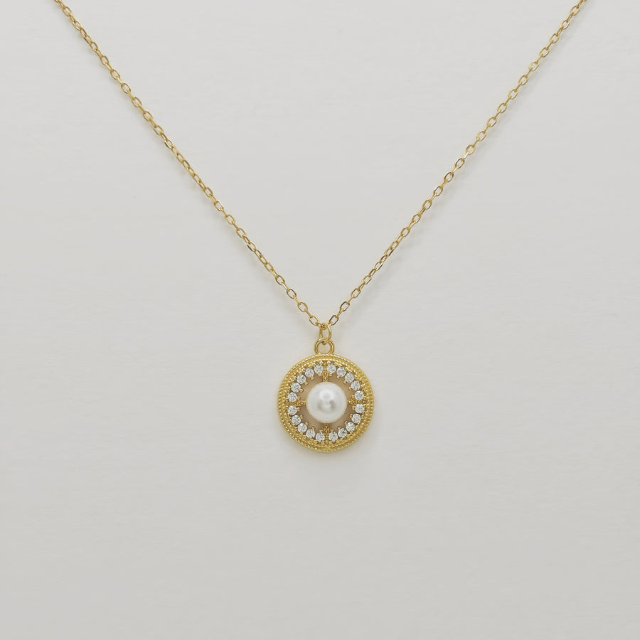 This elegant necklace features a delicate gold chain and a round pendant. The pendant showcases a central pearl surrounded by a circle of small, sparkling cubic zircon, adding a touch of sophistication and luxury. Perfect for both special occasions and everyday wear, this necklace combines timeless beauty with modern elegance.