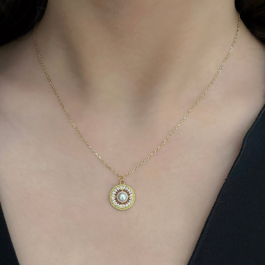 This elegant necklace features a delicate gold chain and a round pendant. The pendant showcases a central pearl surrounded by a circle of small, sparkling cubic zircon, adding a touch of sophistication and luxury. Perfect for both special occasions and everyday wear, this necklace combines timeless beauty with modern elegance.