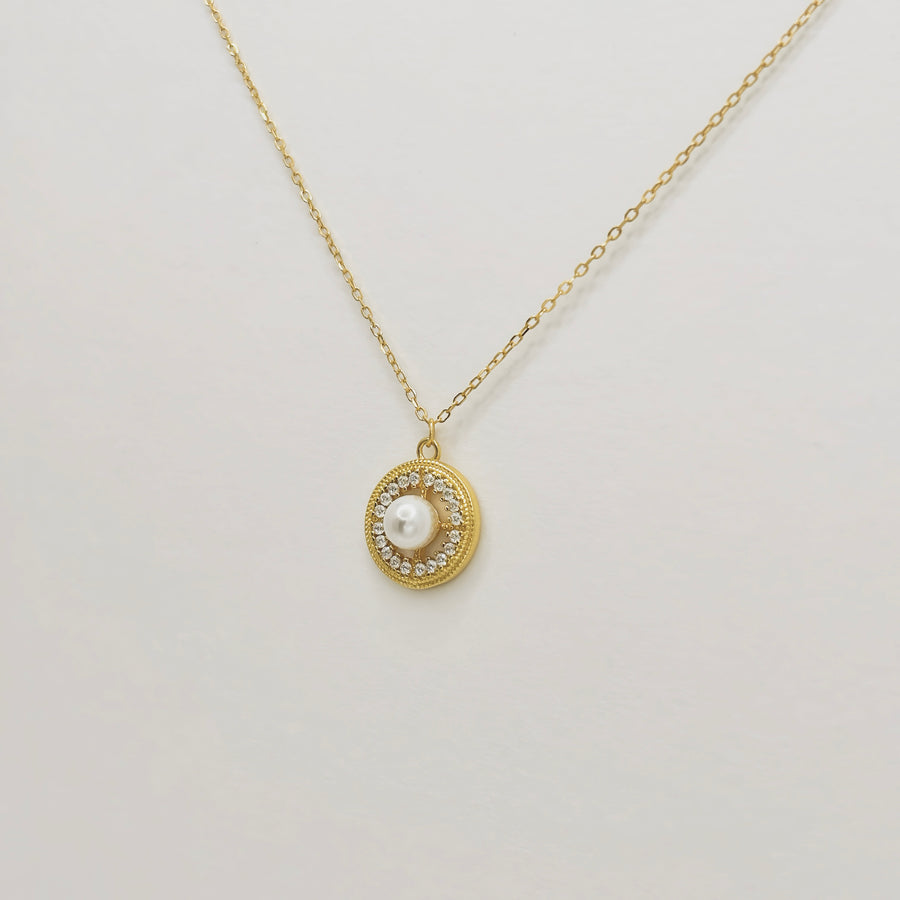 This elegant necklace features a delicate gold chain and a round pendant. The pendant showcases a central pearl surrounded by a circle of small, sparkling cubic zircon, adding a touch of sophistication and luxury. Perfect for both special occasions and everyday wear, this necklace combines timeless beauty with modern elegance.