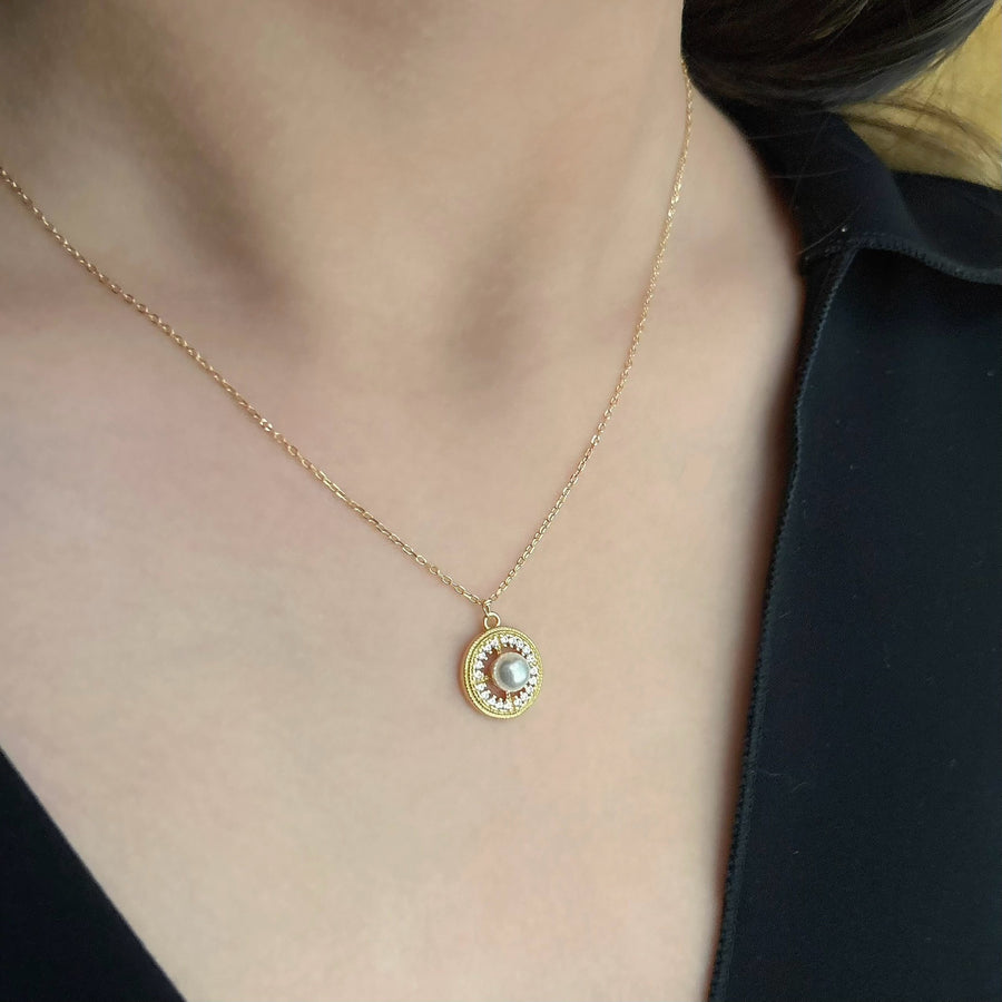 This elegant necklace features a delicate gold chain and a round pendant. The pendant showcases a central pearl surrounded by a circle of small, sparkling cubic zircon, adding a touch of sophistication and luxury. Perfect for both special occasions and everyday wear, this necklace combines timeless beauty with modern elegance.