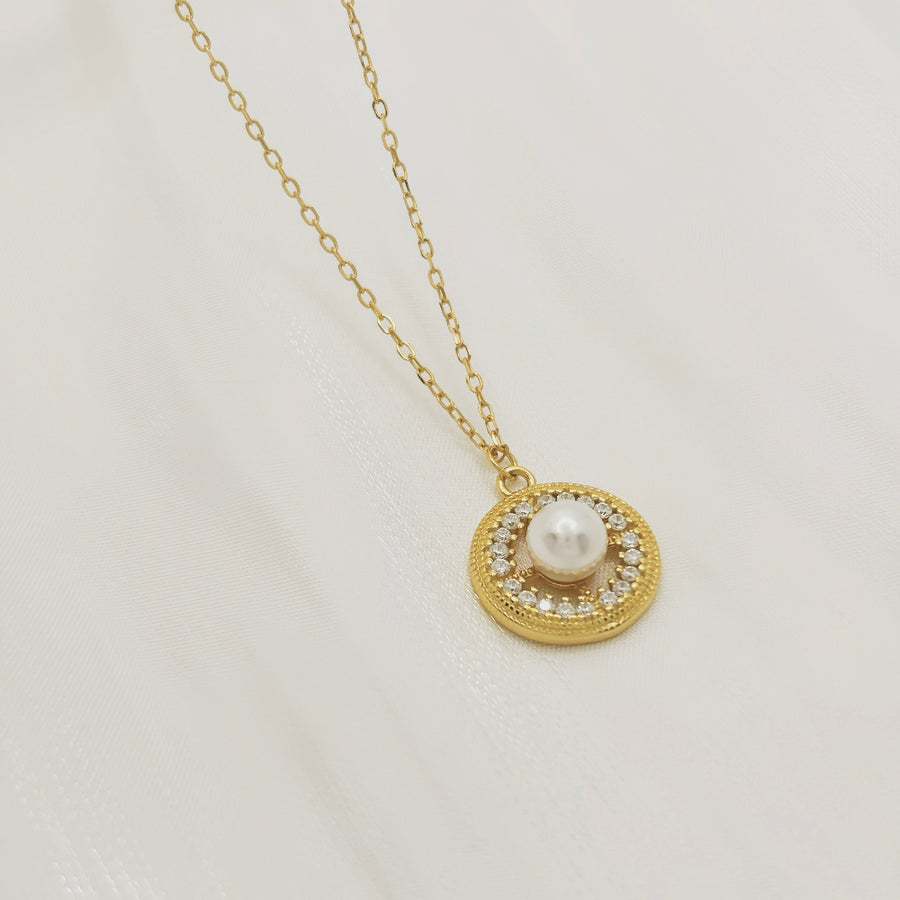 This elegant necklace features a delicate gold chain and a round pendant. The pendant showcases a central pearl surrounded by a circle of small, sparkling cubic zircon, adding a touch of sophistication and luxury. Perfect for both special occasions and everyday wear, this necklace combines timeless beauty with modern elegance.