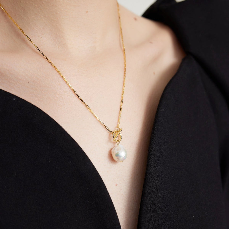 This necklace features a large, irregularly shaped baroque pearl pendant suspended from a gold chain. The chain has a toggle clasp with a circular ring and a horizontal bar, adding a stylish and functional element to the design. Perfect for both special occasions and everyday wear, this necklace combines the natural beauty of baroque pearls with contemporary elegance.